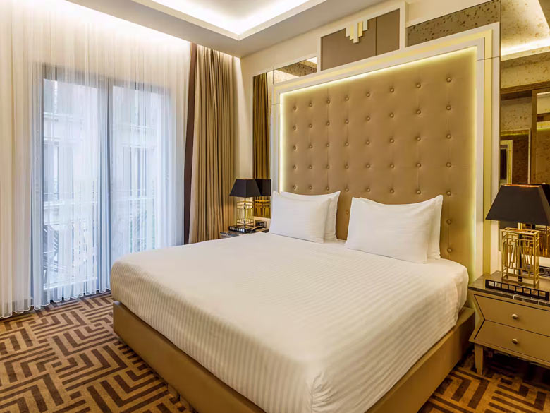 A luxuriously furnished hotel room with white and gold details. A large bed is located in the center.