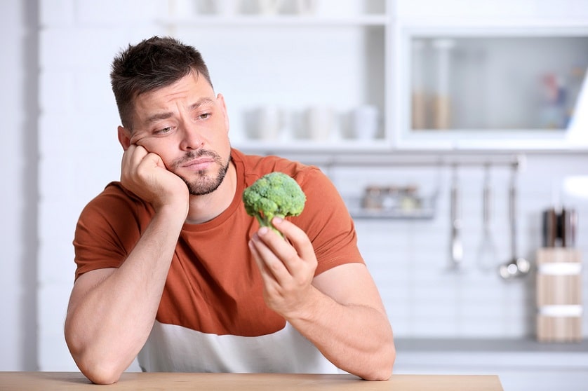Mann hält ein Stück Brokkoli in der Hand während er darüber nachdenkt ob die vegane Ernährung seine Haare ausfallen lässt