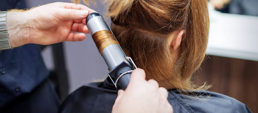 Frau ist beim Friseur, welcher ihre Haare mit einem Lockenstab frisiert