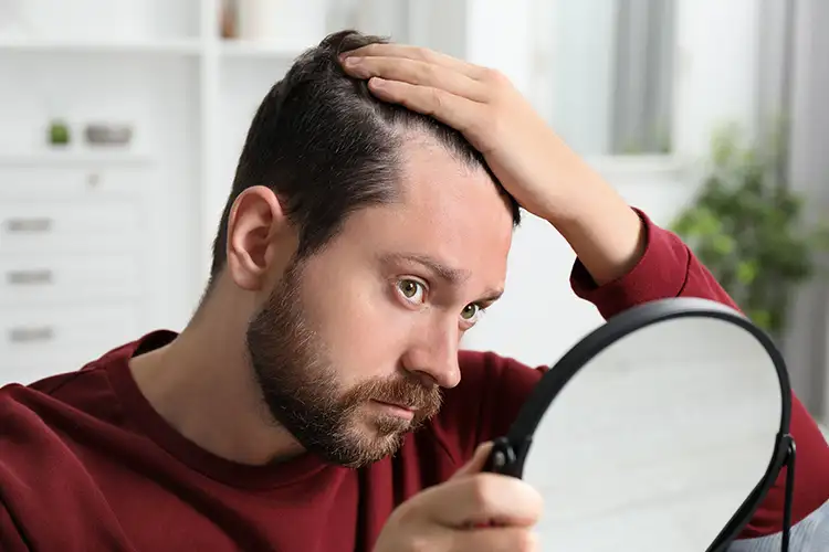 Ein Mann zieht sich mit einer Hand die Haare zur Seite und schaut dabei seine Geheimratsecken im Spiegel an.
