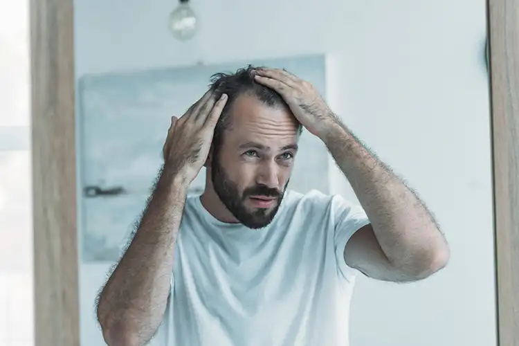 Ein Mann steht vor dem Spiegel und fasst sich mit beiden Händen durch sein Haar. Man erkennt Geheimratsecken und er schaut unglücklich aus.