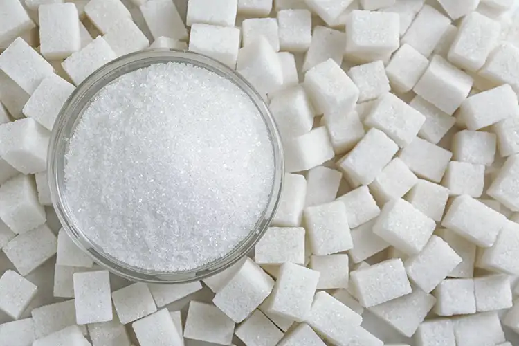 Viele Zuckerwürfel liegen nebeneinander. Links auf den Würfeln steht eine Schale mit losem Zucker.